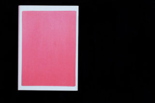 Ein geschlossenes Buch mit schlichtem roten Einband und weißem Rand steht vor einem schwarzen Hintergrund.