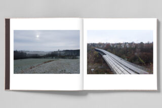Ein aufgeschlagenes Buch zeigt zwei Landschaftsfotos: links ein nebliges Feld und rechts eine geschwungene Fußgängerbrücke, die zu den Häusern führt.
