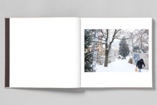 Auf einer Seite des Fotobuchs ist eine verschneite Stadtszene mit einer gehenden Person, Bäumen, einer mit Graffiti bedeckten Wand und einem Mast mit Plakaten zu sehen.