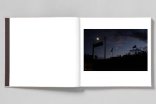 Ein auf einer Seite aufgeschlagenes Fotobuch, das eine Nachtszene mit einer Straßenlaterne, Flaggen, einem Wegweiser und dem Mond in einem dunklen, teilweise bewölkten Himmel zeigt.