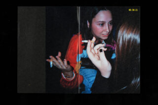 Zwei Personen sitzen in einem dunklen Raum eng beieinander. Eine gestikuliert und hält eine Zigarette in der Hand; die andere hört zu und hält ebenfalls eine Zigarette in der Hand. In der Ecke erscheint ein digitaler Zeitstempel.