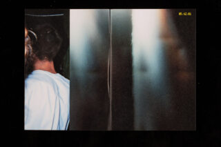 Links steht eine Person in weißem Hemd mit dem Rücken zur Kamera; rechts ist eine abstrakte, verschwommene vertikale Spiegelung zu sehen. Oben rechts ist ein gelber Zeitstempel mit der Aufschrift „05·12·01“ zu sehen.