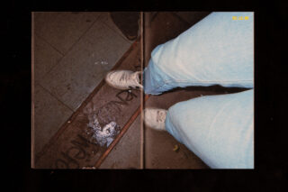 Eine Person in hellblauen Jeans und weißen Turnschuhen steht auf einem gefliesten Boden neben einer Fußmatte mit teilweise sichtbaren Buchstaben.