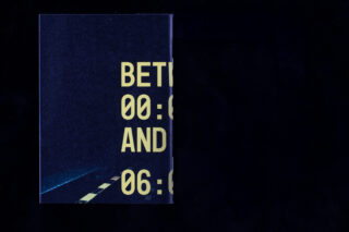Ein dunkelblaues Schild, das teilweise vor einem schwarzen Hintergrund sichtbar ist und großen gelben Text mit Uhrzeiten zeigt: „BET… 00:… AND 06:…“ sowie einen Teil einer Straßengrafik.