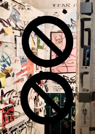 Eine Toilettenkabine voller Graffiti, Aufkleber und Poster, über die zwei große Verbotssymbole gelegt sind.