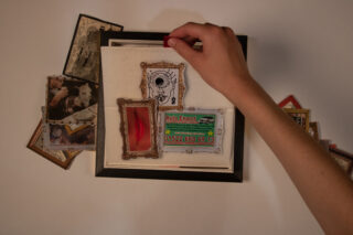 Eine Hand arrangiert kleine gerahmte Fotos, eine Visitenkarte und eine dekorative Karte auf einer weißen Oberfläche, umgeben von verstreuten Vintage-Fotos.