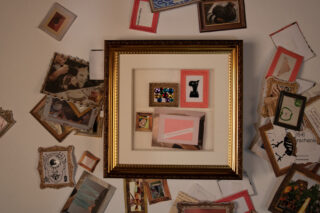 Ein gerahmtes Kunstwerk mit verschiedenen kleinen Objekten und Bildern steht zentriert auf einer weißen Fläche, umgeben von verstreuten Fotografien und Kunstdrucken in unterschiedlichen Rahmen.
