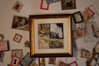 Eine gerahmte Schwarzweißillustration steht mittig an einer Wand, umgeben von vielen kleinen, verstreuten Bilderrahmen mit Fotos, Zeichnungen und ausgeschnittenem Text.