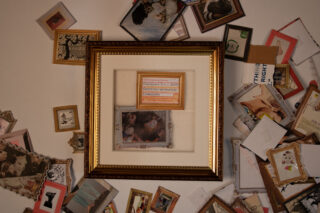 Ein gerahmtes Bild mit einer Zugfahrkarte und einem Foto steht mittig auf einem Tisch, umgeben von verschiedenen verstreuten gerahmten und ungerahmten Fotos und Kunstwerken.