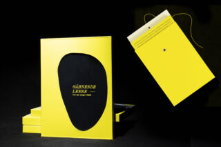 Ein gelbes Buch mit einem unregelmäßigen schwarzen Ausschnitt auf dem Einband und dem Titel „Gähnende Leere“ liegt neben einem gelben Umschlag mit Bindfadenverschluss, beide auf schwarzem Hintergrund.