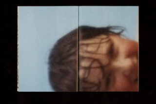 Eine verschwommene Nahaufnahme des nassen Gesichts eines Kindes, das teilweise vor einem blauen Hintergrund sichtbar ist und über die Mittelfalz eines Buches gedruckt ist.