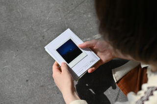 Eine Person hält einen Stapel Sofortbilder in der Hand und betrachtet ein dunkles, verschwommenes Foto, während sie auf einer Betonfläche sitzt.
