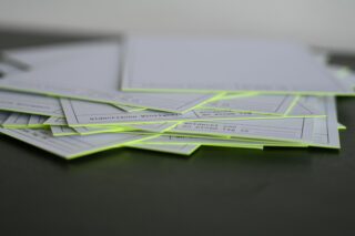 Auf einer dunklen Fläche liegt ein Stapel weißer Karten mit fluoreszierend gelben Rändern. Auf den Karten ist teilweise gedruckter Text zu sehen.