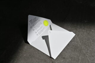 Ein weißer, teilweise geöffneter Papierumschlag mit gedruckten Anweisungen und einem gelben Aufkleber, der auf einer dunklen, strukturierten Oberfläche liegt.