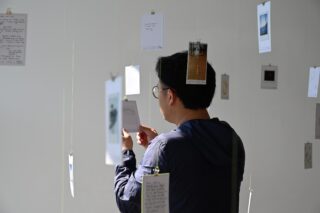 Eine Person mit Brille betrachtet in einem hell erleuchteten Raum an Schnüren hängende Karten und Fotos.