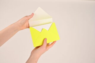 Eine Person hält einen leuchtend gelben Umschlag mit einer teilweise eingelegten weißen Karte auf neutralem Hintergrund.