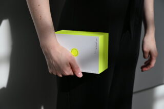 Eine Person in schwarzer Kleidung hält ein dickes, weiß-neongelbes Buch mit minimalistischem Design an ihre Seite.