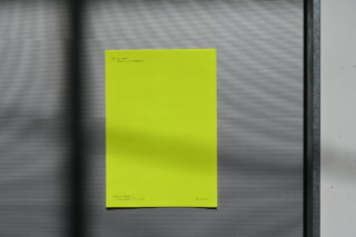 Ein leuchtend gelbes Blatt Papier mit klein gedrucktem Text ist auf einer grauen, gerippten Oberfläche mit horizontalen Linien und Schatten befestigt.