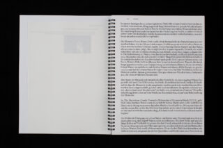Ein spiralgebundenes Buch ist geöffnet. Es zeigt eine leere linke Seite mit einem schwachen Bild und eine rechte Seite, die dicht mit deutschem Text gefüllt ist.