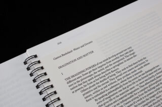 Eine Nahaufnahme eines offenen Spiralbuchs, das die erste Seite eines Kapitels mit dem Titel „Imagination und Materie“ aus „Wasser und Träume“ von Gaston Bachelard zeigt.