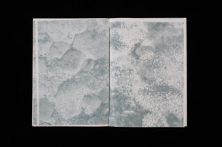 Ein offenes Buch zeigt zwei Seiten mit abstrakten, strukturierten blaugrauen Mustern, die Nahaufnahmen organischer oder geologischer Oberflächen ähneln.