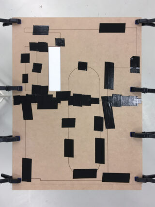 Eine braune Tafel mit aufgedrucktem Umriss und mehreren schwarzen Klebebandrechtecken, einem weißen Rechteck und vier Klammern, die die Tafel an den Rändern festhalten.