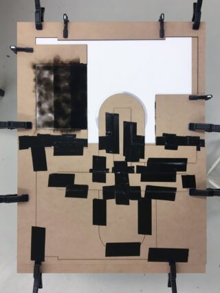 Eine rechteckige Platte ist an den Rändern festgeklemmt und mit schwarzem Klebeband sowie einem rauchähnlichen Muster auf weißem Papier versehen, das in geometrischen Formen und Linien angeordnet ist.
