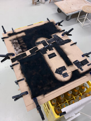 Ein Holzbrett ist an einem Tisch befestigt und mit schwarzer Sprühfarbe und Klebeband bedeckt. Darunter sind gelbe Flaschenkisten und im Hintergrund Hocker zu sehen.