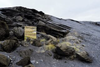 Felsiger, grauer Hang mit geschichteten Formationen und spärlichem gelbem Gras; ein rechteckiger Spiegel reflektiert eine helle, grasbewachsene Landschaft mit einem Bach.