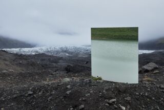 Auf felsigem Boden in der Nähe eines Gletschers steht ein großer Spiegel, der einen grünen Hügel und Wasser reflektiert, das in der unmittelbaren Umgebung nicht sichtbar ist.