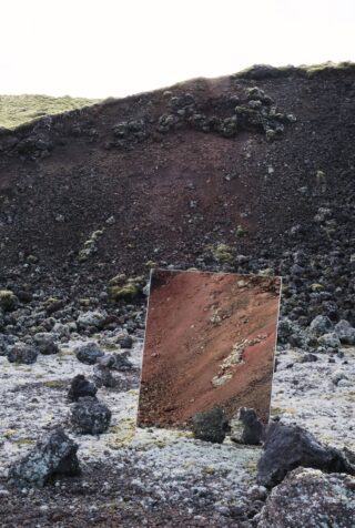 Ein rechteckiger Spiegel ist zwischen dunklem Vulkangestein platziert und reflektiert einen rötlichen Fleck Erde, der mit der felsigen Landschaft im Hintergrund verschmilzt.