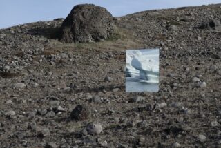 Ein großer Felsen liegt auf einem felsigen, kargen Hang. Im Vordergrund reflektiert ein rechteckiger Spiegel eine eisige Gletscherszene, die einen Kontrast zur trockenen Landschaft bildet.