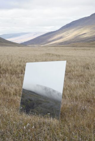 Ein rechteckiger Spiegel steht aufrecht auf einer Wiese und reflektiert einen nebligen Hügel und Himmel, der einen Kontrast zur trockenen Landschaft um ihn herum bildet.
