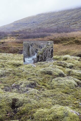 Auf moosigem Boden in einer Graslandschaft steht ein Spiegel, der das Bild eines felsigen Wasserfalls reflektiert, der einen Kontrast zum umgebenden Gelände bildet.