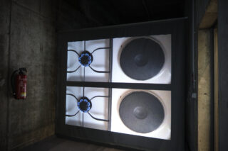 Zwei Bilder eines Gasherdbrenners und einer Elektroherdspule werden nebeneinander auf eine Wand in einem Industriegebäude projiziert. In der Nähe ist ein Feuerlöscher angebracht.