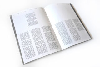 Auf einer weißen Fläche liegt ein offenes Buch, das zwei Seiten schwarz gedruckten Text enthält, der in drei Spalten pro Seite angeordnet ist.