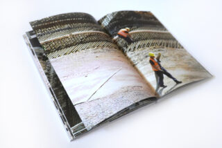 Offenes Buch mit Fotos einer Baustelle, auf der Arbeiter in orangefarbenen Westen und gelben Helmen auf einer Betonoberfläche stehen, auf der Stahlbewehrungen sichtbar sind.