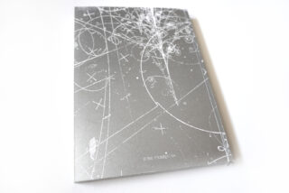 Ein silbernes Buch mit weißen geometrischen und abstrakten Linienmustern auf dem Cover, unten in der Mitte mit dem Titel „JUNE FABREGAS“, fotografiert vor einem weißen Hintergrund.