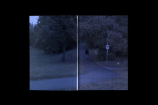 Eine Person geht in der Abenddämmerung allein auf einem gepflasterten Weg durch einen Park, umgeben von Gras und Bäumen. Ein Wegweiser und ein Spalt verlaufen vertikal durch das Bild.