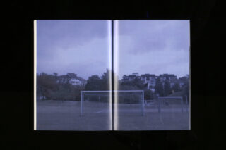 Ein offenes Buch mit dem Foto eines Fußballfelds mit zwei leeren Torpfosten, Bäumen und Gebäuden im Hintergrund unter einem bewölkten Himmel.
