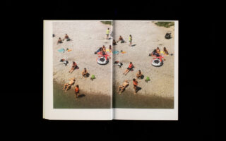 Ein offenes Buch mit einer Luftaufnahme von Menschen, die mit Handtüchern und Sonnenschirmen an einem felsigen Strand sonnenbaden und faulenzen.