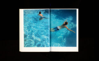 In einem offenen Buch ist ein Foto von zwei Personen zu sehen, die in einem klaren, blauen Pool schwimmen. Auf gegenüberliegenden Seiten schwimmen sie jeweils in entgegengesetzte Richtungen.