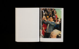 Ein offenes Buch mit dem Foto eines Mannes, der in einer überfüllten Umgebung im Freien in der Nähe von Wasser ein Selfie macht.