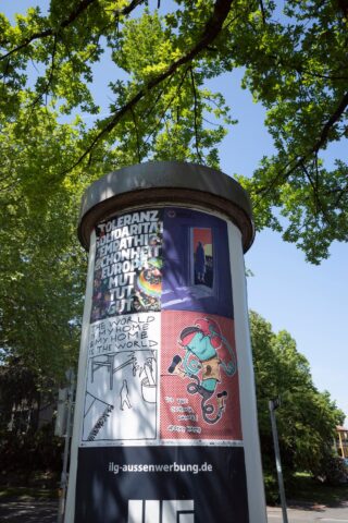 Eine zylindrische Litfaßsäule im Freien zeigt Plakate, die für Toleranz, Einheit und Kunstveranstaltungen werben, teilweise im Schatten grüner Äste.
