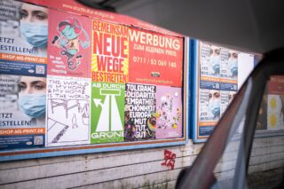 Eine Reihe farbenfroher Plakate mit deutschem Text, Grafiken und Anzeigen sind auf einer Plakatwand an der Straße zu sehen und teilweise durch ein Autofenster zu sehen.