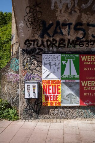 Eine verwitterte Wand mit Graffiti und vier Plakaten, neben einem kleinen Verkaufsautomaten, auf einem sonnenbeschienenen Gehweg mit Grün im Hintergrund.
