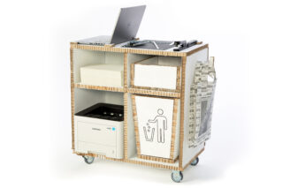 Ein mobiler Arbeitsplatz mit Laptop, Drucker, Papierfächern, Recyclingbehälter und Zeitungshalter, alles untergebracht in einem Wagen aus Pappe und Holz auf Rädern.