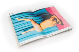 Auf einer aufgeschlagenen Zeitschrift ist das Foto einer nackten Person zu sehen, die auf der Seite liegt, vor einem blau-rosa Hintergrund.