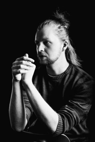 Schwarz-Weiß-Porträt einer Person mit zurückgebundenen langen Haaren, die einen Pullover trägt, mit gefalteten Händen dasitzt und nachdenklich zur Seite blickt.