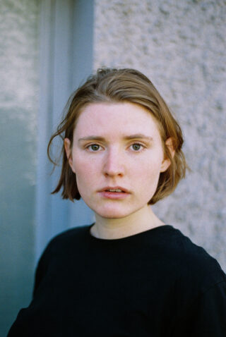 Eine Person mit kurzen, hellbraunen Haaren und heller Haut steht vor einer strukturierten Wand, trägt ein schwarzes Hemd und blickt mit neutralem Gesichtsausdruck direkt in die Kamera.